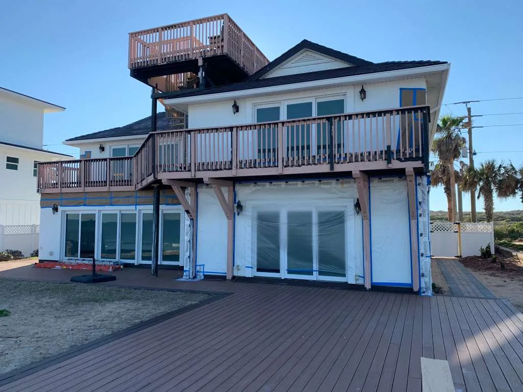 An oceanfront home before, with bare wood deck railings and windows masked for prep