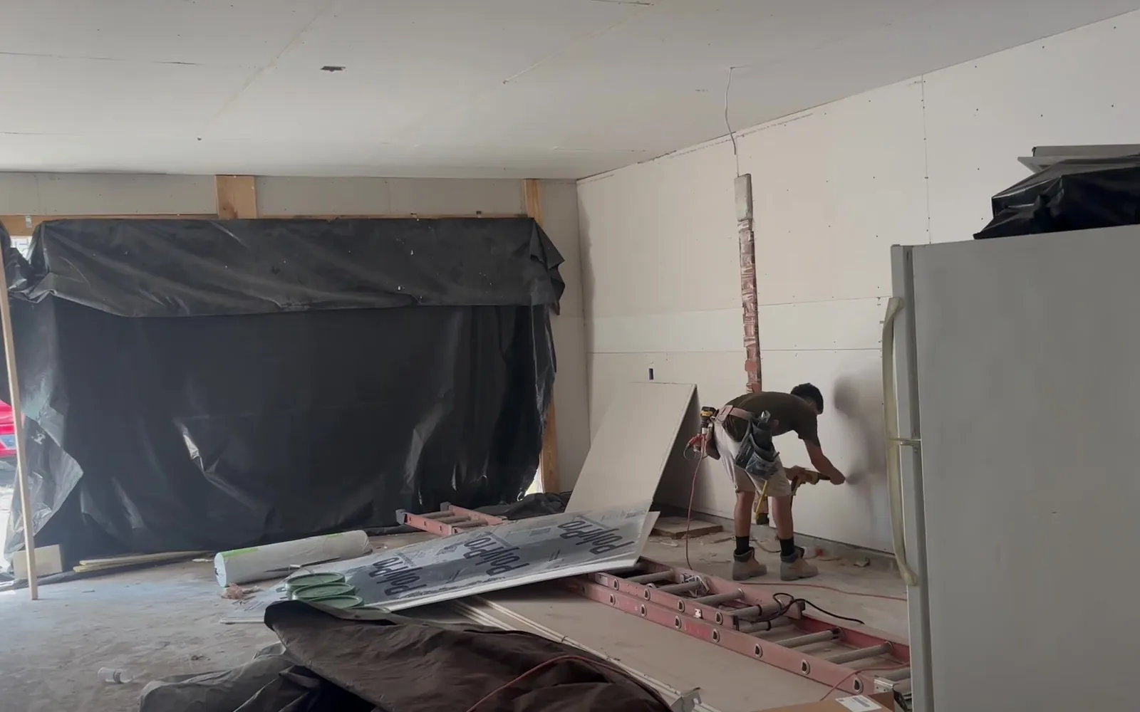A crew member fastening a new sheet of drywall while furniture stays covered in plastic
