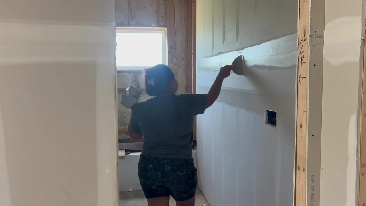 A crew member knifing a drywall seam smooth beside a window